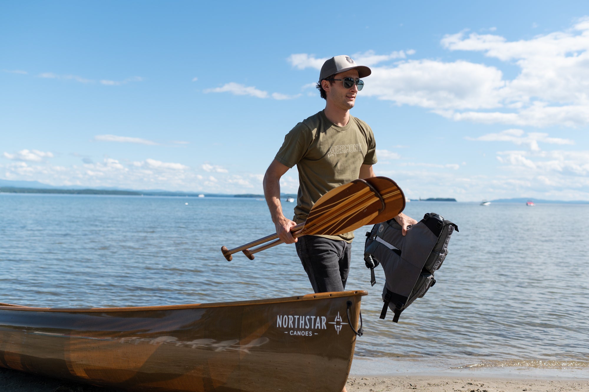 Person carrying two canoe paddles and a life jacket past a canoe on a lake beach in Vermont wearing a Vermont Eclectic Company t-shirt