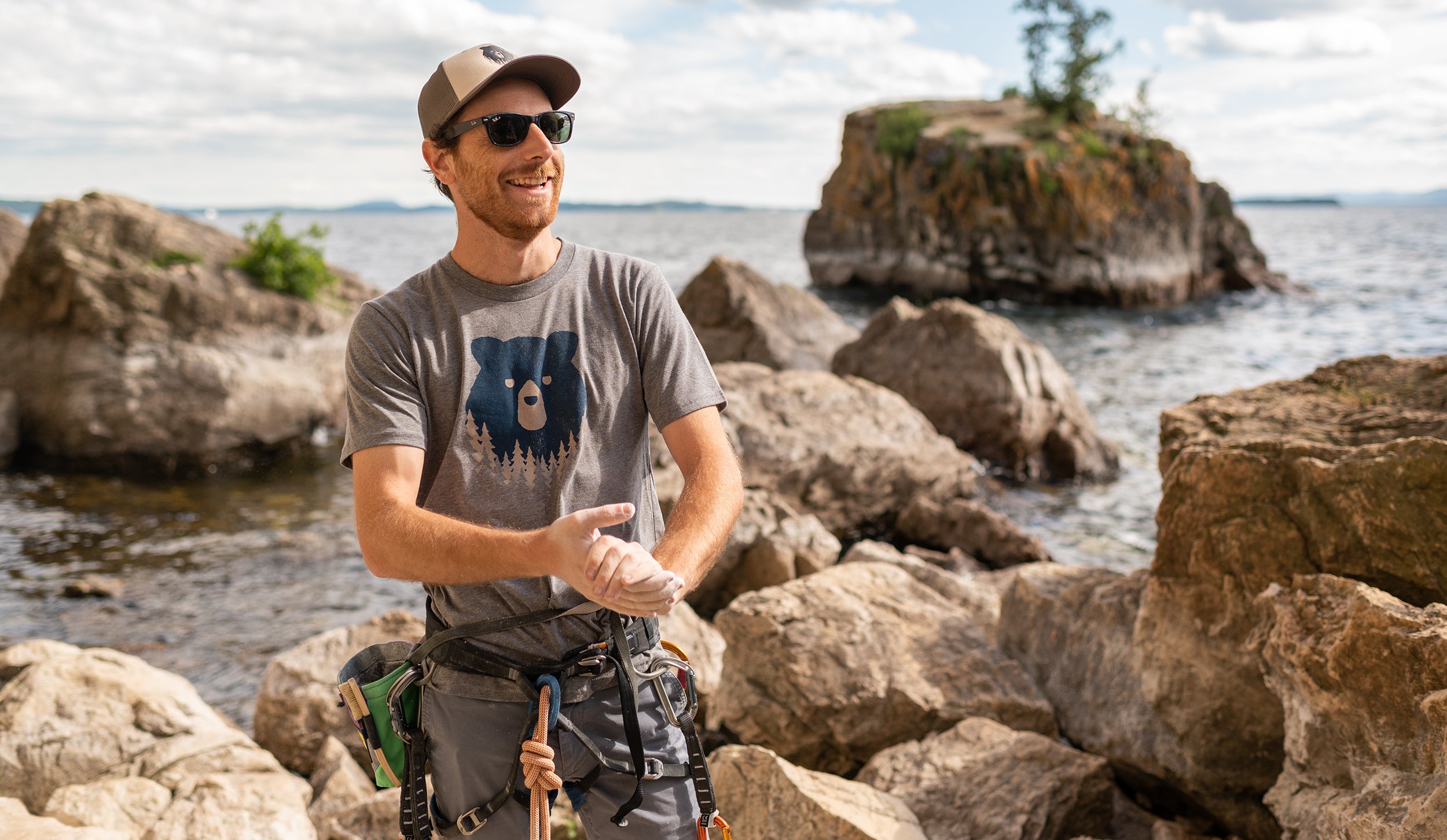 Person in Vermont Eclectic Company t-shirt with rock climbing gear standing on boulders in front of water