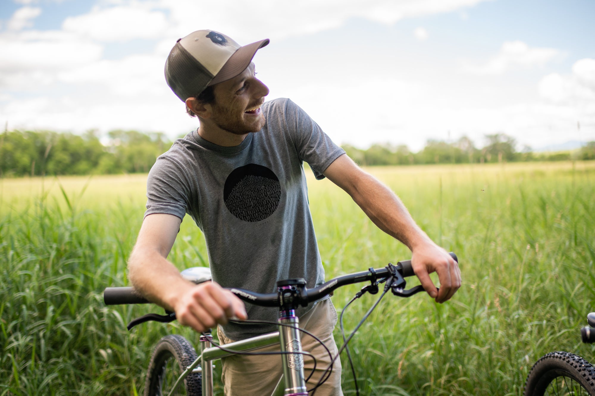 Man wearing Vermont Eclectic Company t-shirt and trucker hat leaning on his mountain bike in a green field