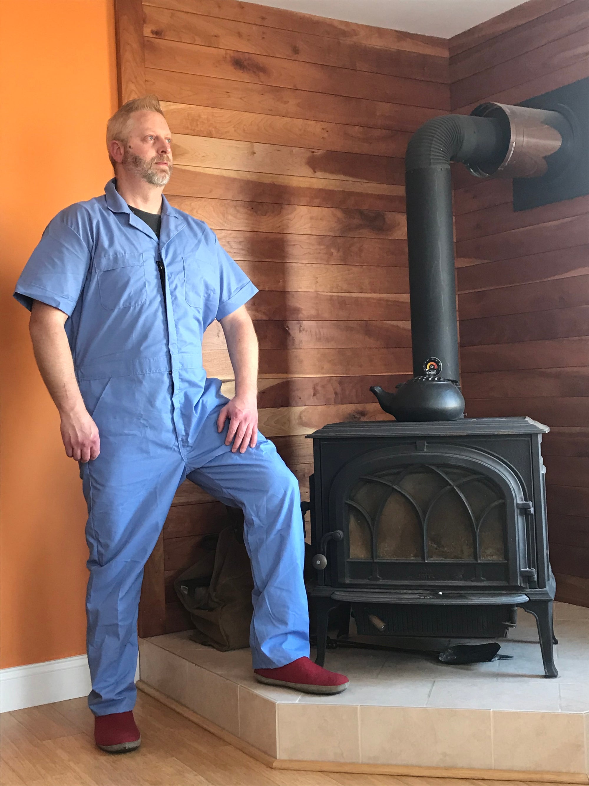 David Holub in a blue jumpsuit standing in front of a wood burning stove