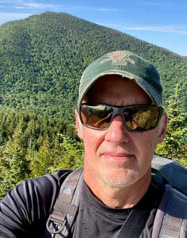 Todd Cummings in a green hat and sunglasses with a green mountain in the background