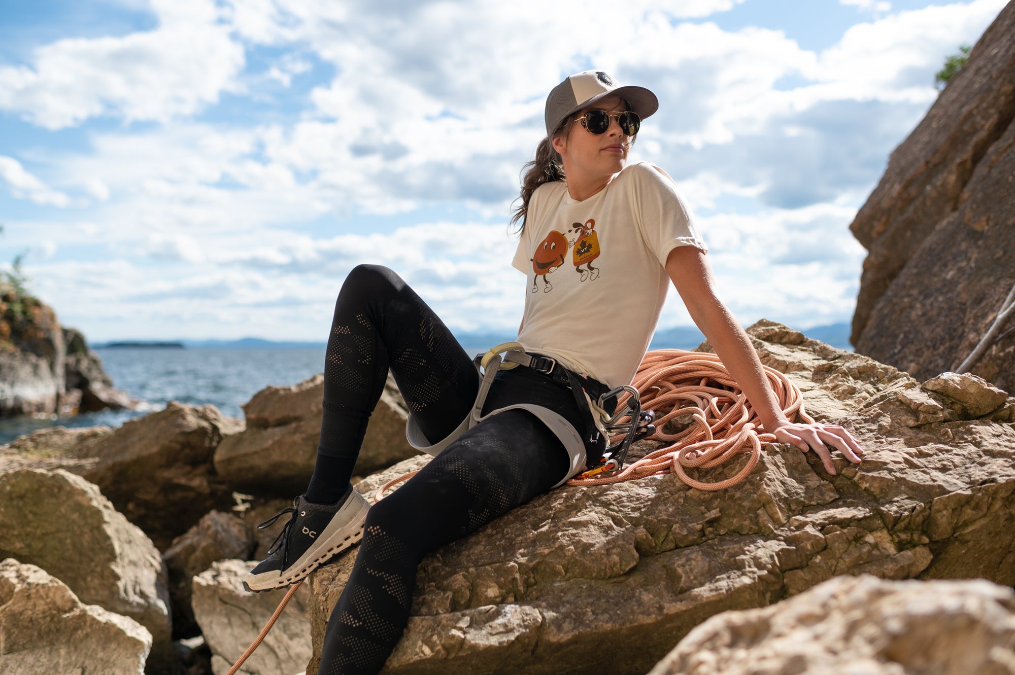 Woman sitting on rocks with climbing gear, wearing a Vermont Eclectic Company trucker hat and t-shirt