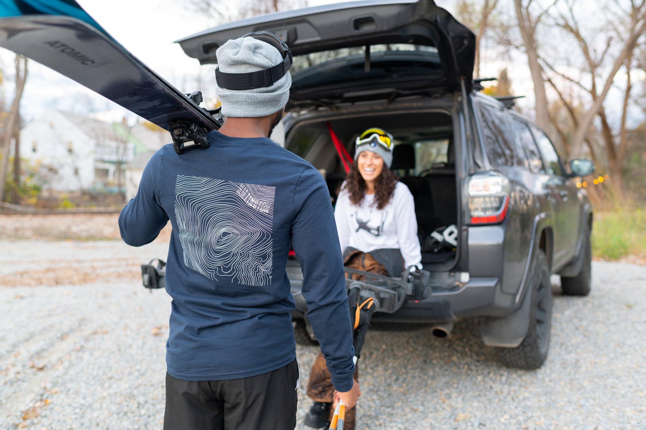 Person carrying skis towards a car with a person sitting on the back of car both wearing Vermont Eclectic Company t-shirts